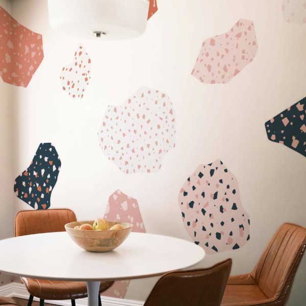 A modern abstract wallpaper design with floating terrazzo-like shapes in soft pink, coral, and navy tones scattered across a creamy beige background. Set in a cozy dining nook with caramel leather chairs and a round white table, the mural adds a playful yet elegant touch. The Fancy Walls peel and stick or non-pasted wallpaper is available in custom sizes for your wall.