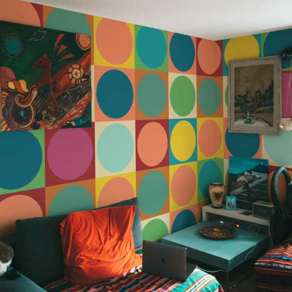 A vivid geometric mural filled with oversized colorful circles in bright hues like teal, orange, pink, and yellow, arranged in a bold grid. Shown in a lively living room with eclectic decor, patterned fabrics, and modern art, this mural energizes the space with retro charm. The Fancy Walls peel and stick or non-pasted wallpaper is available in custom sizes for your wall.