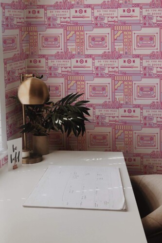 Modern home office decorated with pink 2000s pattern wallpaper featuring bold, retro illustrations of cassette tapes, media players, and nostalgic text like “Do you miss in 2000s?” in purple and magenta outlines. The accent wall adds a fun Y2K vibe to the workspace, which includes a sleek white desk, brass table lamp, leafy potted plant, and a motivational “be bold” card. Soft natural light from the window enhances the playful, creative setting.