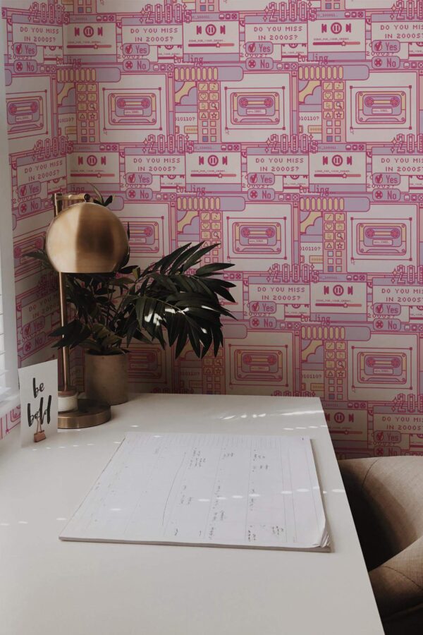 Modern home office decorated with pink 2000s pattern wallpaper featuring bold, retro illustrations of cassette tapes, media players, and nostalgic text like “Do you miss in 2000s?” in purple and magenta outlines. The accent wall adds a fun Y2K vibe to the workspace, which includes a sleek white desk, brass table lamp, leafy potted plant, and a motivational “be bold” card. Soft natural light from the window enhances the playful, creative setting.