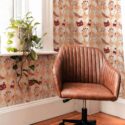 A warm, eclectic nook showcasing an Aesthetic Witch Halloween wallpaper with burgundy cat masks and hand motifs on a beige-and-pale-pink diamond background, paired with a brown leather swivel chair, wooden floors, and a potted plant by the window.