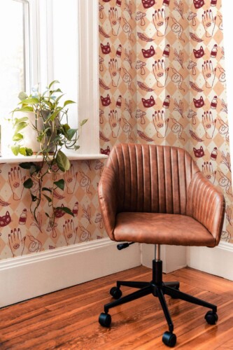 A warm, eclectic nook showcasing an Aesthetic Witch Halloween wallpaper with burgundy cat masks and hand motifs on a beige-and-pale-pink diamond background, paired with a brown leather swivel chair, wooden floors, and a potted plant by the window.