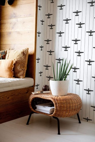 Airplane Wallpaper with black plane silhouettes on vertical white stripes, styled in a cozy nook with warm wood paneling, a woven rattan table, neutral cushions, and a potted succulent. The minimalist, monochrome pattern adds playful yet modern energy to the space.