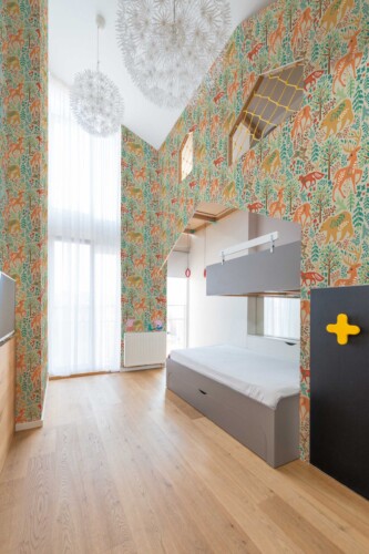 Spacious children’s bedroom with soaring ceilings and large windows draped in sheer white curtains. The tall walls are covered in a colorful woodland wallpaper featuring stylized forest animals—like deer and foxes—among whimsical foliage in shades of teal, orange, and yellow. A gray bunk bed with built-in storage sits against the wall, complemented by natural wood flooring. Modern pendant lights resembling dandelion spheres hang above, creating a bright, playful atmosphere.