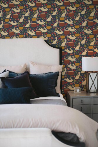 Dark autumn wallpaper with playful rabbits, pumpkins, tree stumps, and berries scattered across a deep navy blue background, adding a cozy and whimsical woodland touch. Displayed in a bedroom with soft bedding and velvet pillows, this design brings warmth and charm to the space. Suitable for peel and stick or non-pasted application, this wallpaper from Fancy Walls is made from PVC-free materials, offering an eco-conscious update for fall-inspired interiors.