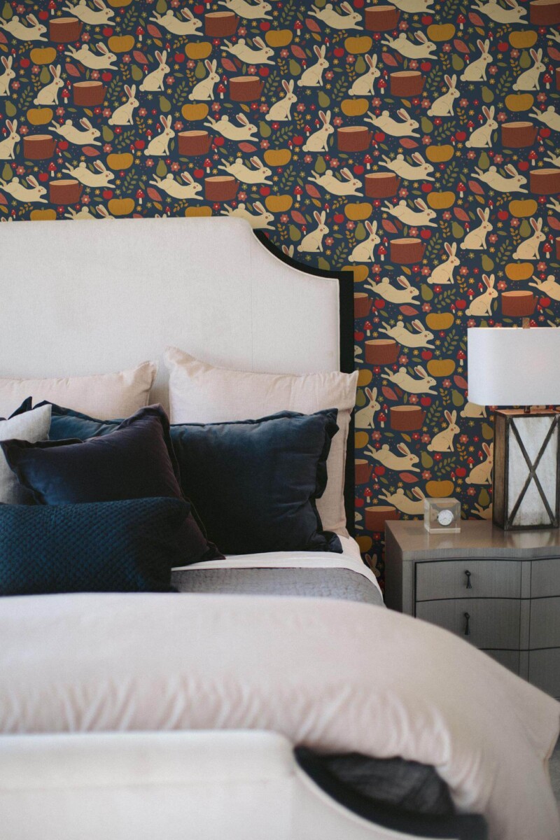 Dark autumn wallpaper with playful rabbits, pumpkins, tree stumps, and berries scattered across a deep navy blue background, adding a cozy and whimsical woodland touch. Displayed in a bedroom with soft bedding and velvet pillows, this design brings warmth and charm to the space. Suitable for peel and stick or non-pasted application, this wallpaper from Fancy Walls is made from PVC-free materials, offering an eco-conscious update for fall-inspired interiors.