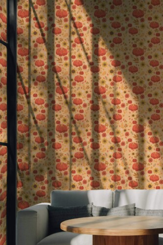 A cozy living room interior featuring a soft beige wall covered in an autumn-inspired wallpaper pattern of orange pumpkins, sunflowers, and red daisies. The hand-drawn motifs are evenly spaced, creating a whimsical fall theme that pairs beautifully with the neutral-toned modern couch and round wooden table. The sun filters in through tall windows, casting artistic shadows that enhance the warmth of the design. The Fancy Walls peel and stick or non-pasted wallpaper adds seasonal charm to living rooms, kitchens, or entryways.