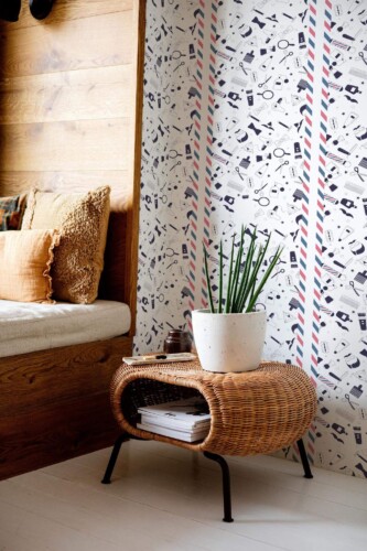 A modern barbershop-inspired wallpaper featuring a playful arrangement of illustrated grooming tools—scissors, combs, razors, hair clippers, and shaving brushes—set against a clean white background with vertical red, white, and blue barber pole stripes. The design adds a fun, novelty character to the space and contrasts with the surrounding natural textures, including a rattan table and soft wood-paneled bench. The Fancy Walls peel and stick or non-pasted wallpaper is ideal for barber shops, salons, or creative studio corners seeking a stylish and lighthearted upgrade.
