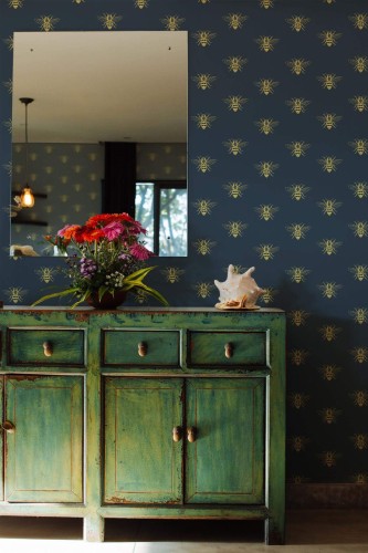 Aesthetic bee wallpaper featuring a repeating pattern of gold-toned bees on a deep navy blue background, styled in an Art Deco-inspired design. The wallpaper adds elegance and visual rhythm to a cozy interior, paired here with a distressed green vintage sideboard, a vibrant floral bouquet, and a seashell for a touch of eclectic charm. The Fancy Walls peel and stick or non-pasted wallpaper is ideal for entryways, living rooms, or accent walls in creative, nature-loving homes.