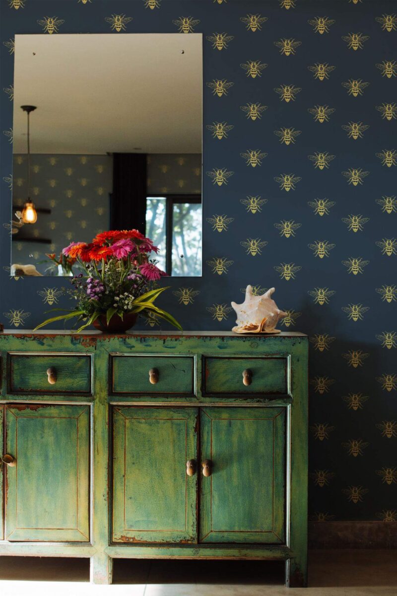 Aesthetic bee wallpaper featuring a repeating pattern of gold-toned bees on a deep navy blue background, styled in an Art Deco-inspired design. The wallpaper adds elegance and visual rhythm to a cozy interior, paired here with a distressed green vintage sideboard, a vibrant floral bouquet, and a seashell for a touch of eclectic charm. The Fancy Walls peel and stick or non-pasted wallpaper is ideal for entryways, living rooms, or accent walls in creative, nature-loving homes.
