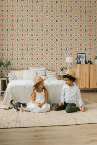 Children’s bedroom featuring a beige retro-style wallpaper covered in repeating round medallion tiles, each filled with colorful illustrations of tropical motifs. The pattern includes orange leopards, blue parrots, palm leaves, and jungle plants arranged in alternating circular frames with dotted borders. A cozy bed with textured white bedding and striped pillows sits against the wall, with two children sitting playfully on a cream woven rug beside a curious kitten. The space feels warm, playful, and nature-inspired.