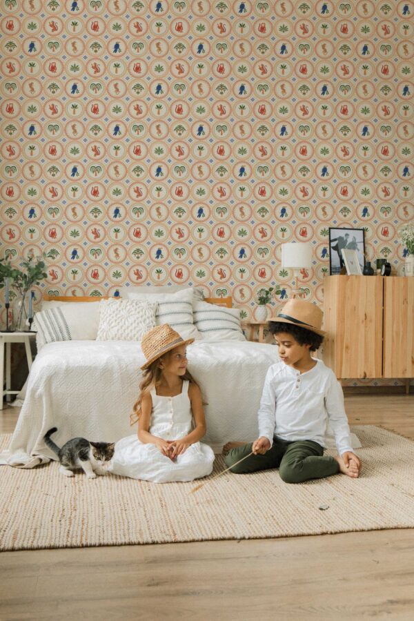 Children’s bedroom featuring a beige retro-style wallpaper covered in repeating round medallion tiles, each filled with colorful illustrations of tropical motifs. The pattern includes orange leopards, blue parrots, palm leaves, and jungle plants arranged in alternating circular frames with dotted borders. A cozy bed with textured white bedding and striped pillows sits against the wall, with two children sitting playfully on a cream woven rug beside a curious kitten. The space feels warm, playful, and nature-inspired.