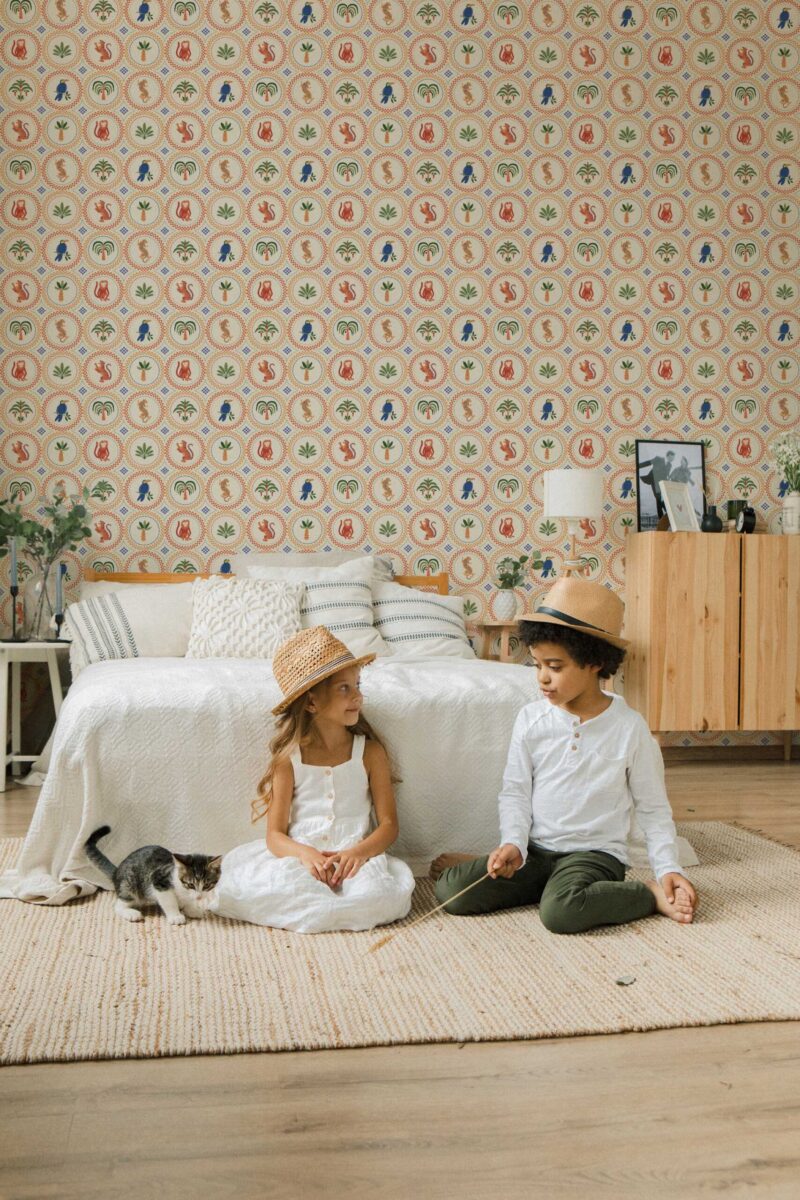 Children’s bedroom featuring a beige retro-style wallpaper covered in repeating round medallion tiles, each filled with colorful illustrations of tropical motifs. The pattern includes orange leopards, blue parrots, palm leaves, and jungle plants arranged in alternating circular frames with dotted borders. A cozy bed with textured white bedding and striped pillows sits against the wall, with two children sitting playfully on a cream woven rug beside a curious kitten. The space feels warm, playful, and nature-inspired.