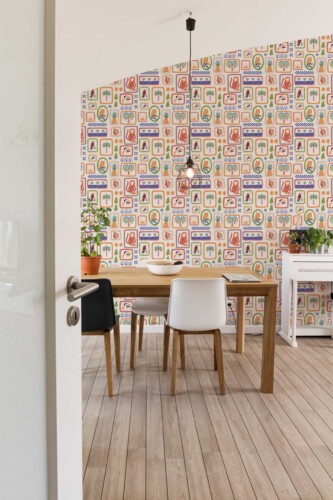 A cozy modern dining room features a statement wall covered in colorful "Cute Fun Tropical" wallpaper by Fancy Walls. The wallpaper showcases a playful grid of tropical-themed frames arranged neatly across a soft beige background. Each rectangular frame contains whimsical illustrations including tigers, monkeys, palm trees, tropical fruits, parrots, papayas, geometric zigzags, and jungle leaves — all painted in vibrant earthy tones of orange, green, red, and blue. The illustrations evoke a hand-drawn, folk-art aesthetic that adds warmth and charm to the space.