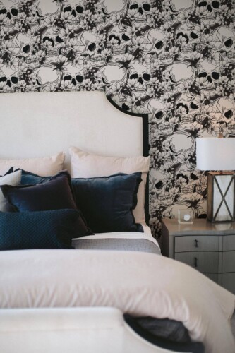 Black and White Skull Wallpaper featuring intricately detailed floral and skull motifs, styled behind a white upholstered bed with plush navy pillows and a gray nightstand holding a lantern-style lamp. The dramatic monochrome pattern adds edgy elegance to the cozy bedroom décor.