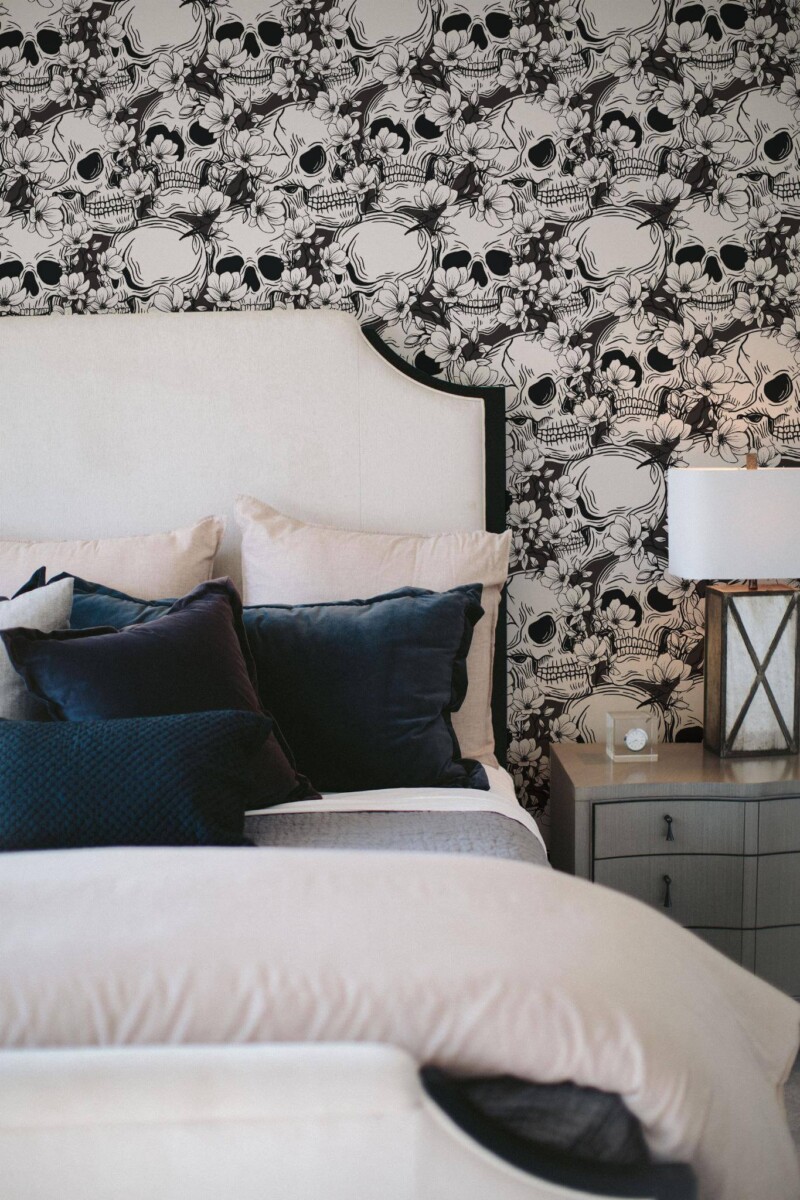 Black and White Skull Wallpaper featuring intricately detailed floral and skull motifs, styled behind a white upholstered bed with plush navy pillows and a gray nightstand holding a lantern-style lamp. The dramatic monochrome pattern adds edgy elegance to the cozy bedroom décor.