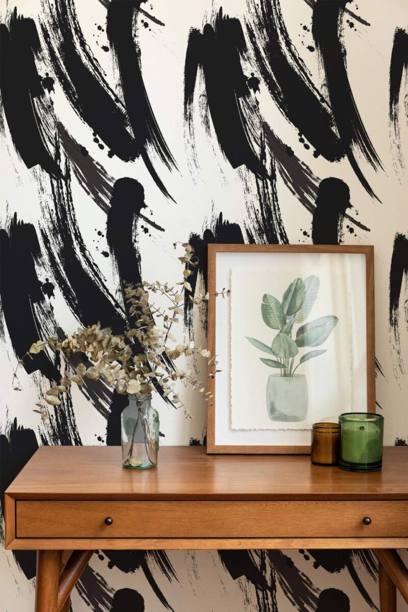 The bold black brush strokes of this wallpaper create a striking, dynamic statement wall in any room. The sweeping strokes, varying in thickness and intensity, evoke an artistic, expressive feel, blending abstract design with a sense of fluid motion. The high contrast of black on a neutral background adds visual depth, making it a perfect fit for modern interiors. This wall features a minimalist, Scandinavian-inspired design with a natural wood table, framed art, and subtle green plants, complementing the wallpaper’s bold artistic theme. The Fancy Walls peel and stick or non-pasted wallpaper brings effortless style and eco-conscious design to your space.