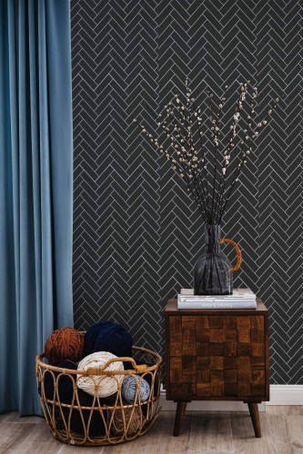 Black Herringbone Wallpaper with a bold, geometric tile pattern on a deep charcoal background, styled in a cozy nook featuring a wooden side table, a teal curtain, and a wicker basket filled with colorful yarn. The crisp lines and dark backdrop add modern sophistication to the space.