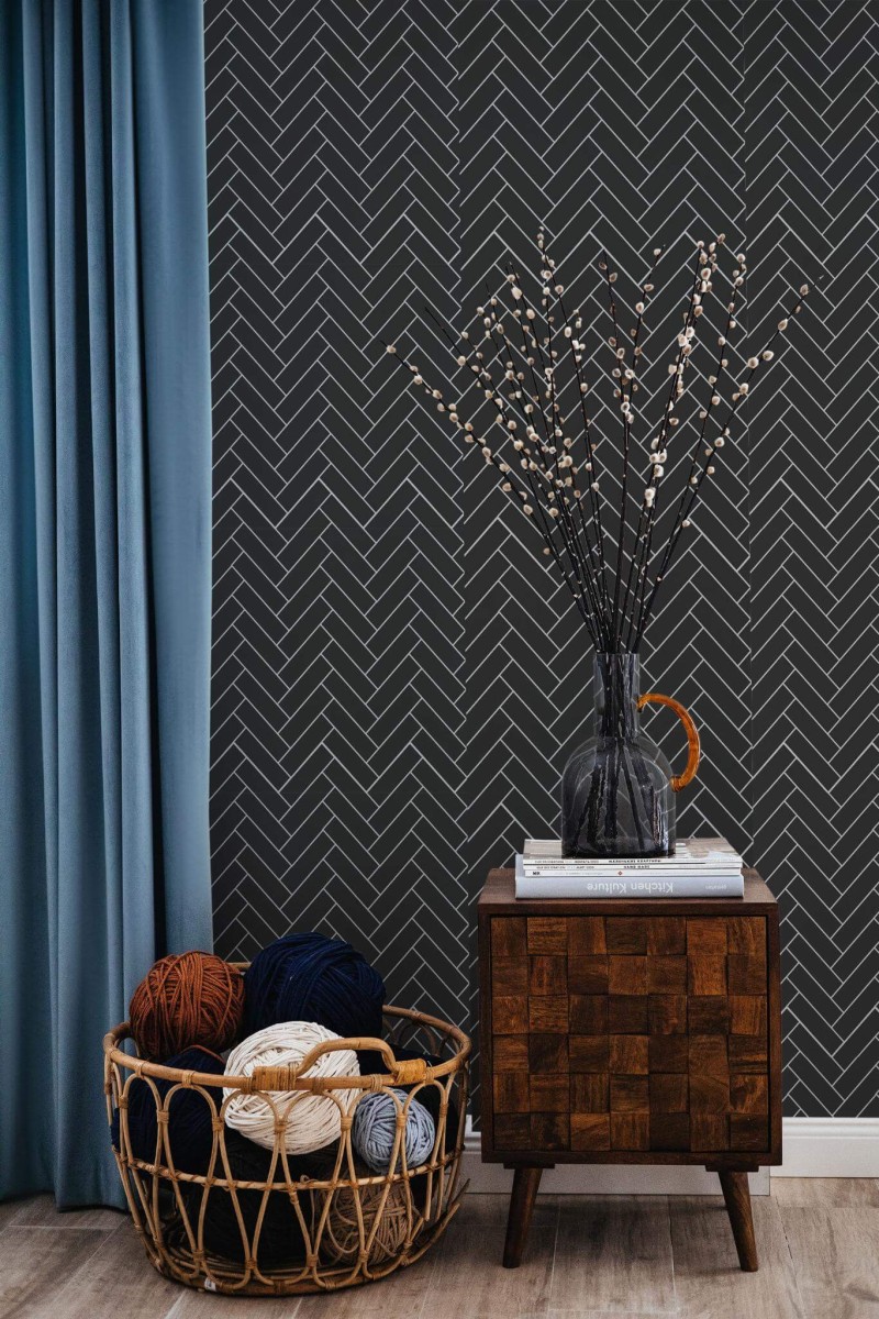 Black Herringbone Wallpaper with a bold, geometric tile pattern on a deep charcoal background, styled in a cozy nook featuring a wooden side table, a teal curtain, and a wicker basket filled with colorful yarn. The crisp lines and dark backdrop add modern sophistication to the space.