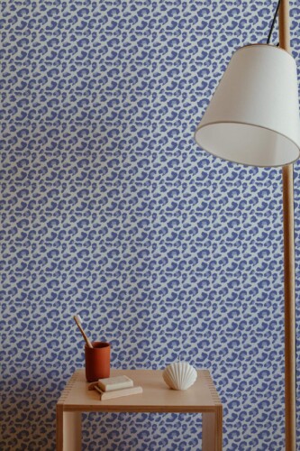 Interior featuring Fancy Walls' Blue Leopard Rhythm wallpaper, a bold and modern animal print design with tightly packed navy and sky-blue leopard spots on a soft cream background. The peel and stick or non-pasted wallpaper adds a playful, contemporary accent to the minimalist setting, which includes a sculptural wood lamp, a light-toned wooden side table, and natural décor items. Ideal for living rooms or reading corners, this eco-conscious design is printed with GreenGuard Gold certified inks.