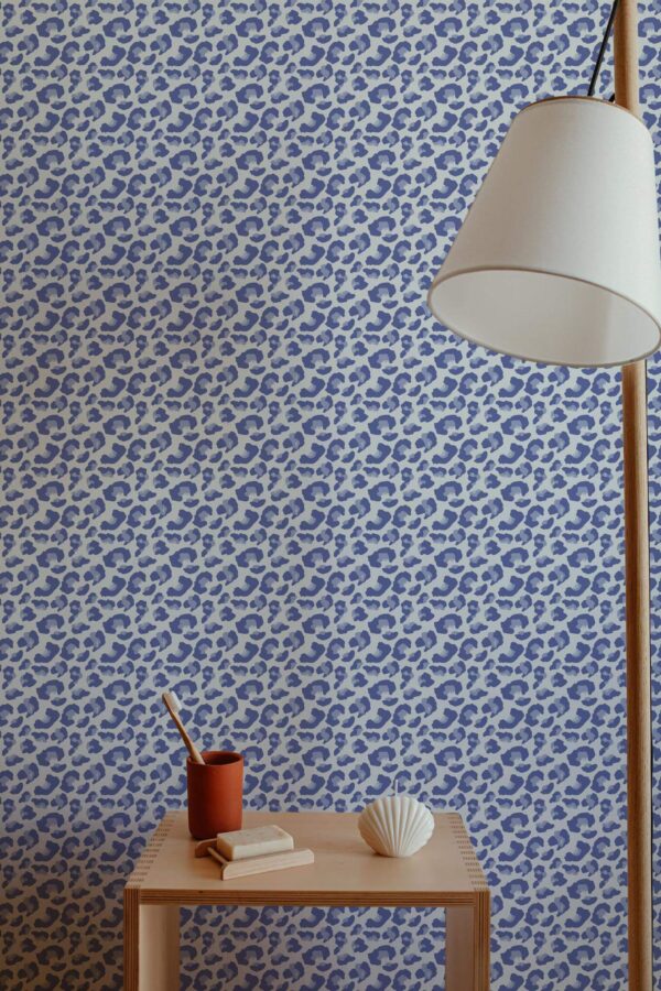 Interior featuring Fancy Walls' Blue Leopard Rhythm wallpaper, a bold and modern animal print design with tightly packed navy and sky-blue leopard spots on a soft cream background. The peel and stick or non-pasted wallpaper adds a playful, contemporary accent to the minimalist setting, which includes a sculptural wood lamp, a light-toned wooden side table, and natural décor items. Ideal for living rooms or reading corners, this eco-conscious design is printed with GreenGuard certified inks.