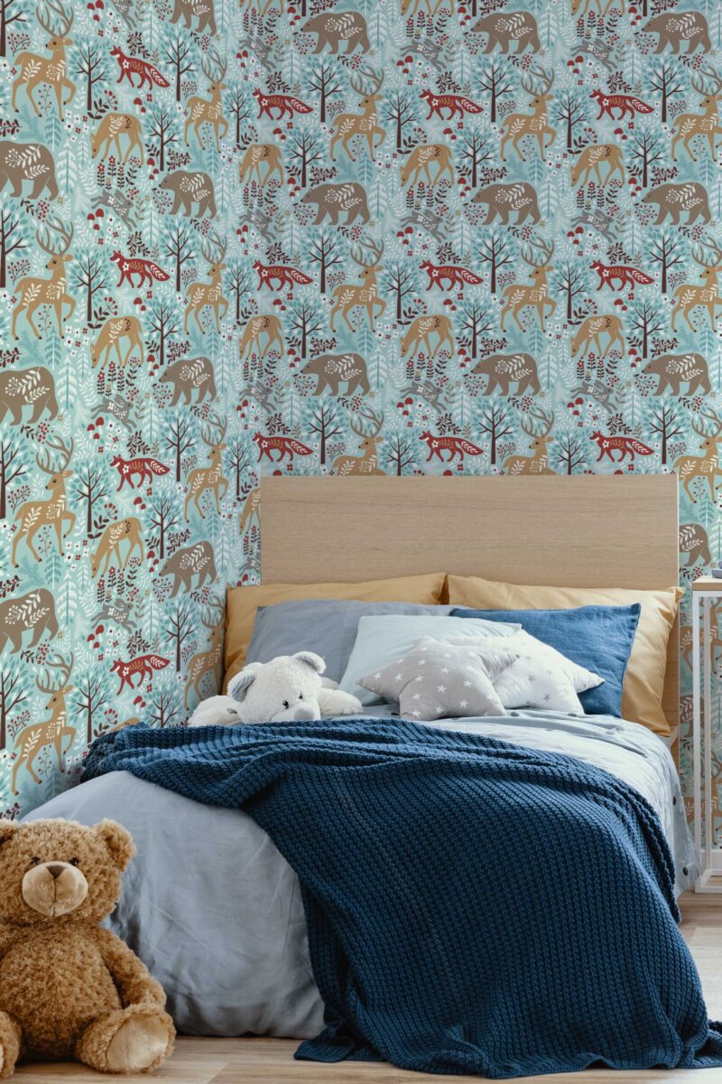Children’s bedroom with a cozy bed draped in a dark blue throw blanket, set against an accent wall featuring a whimsical winter woodland wallpaper. The pattern displays illustrated deer, foxes, and other forest creatures in soft browns and reds, interspersed with blue and white foliage on a pale teal background. Plush teddy bears and pillows add warmth and comfort. The overall scene evokes a calm, storybook ambiance that blends playful animal motifs with soothing winter hues.