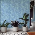 A modern living area featuring a swirling blue marble-patterned wallpaper as the accent wall. The wallpaper design shows flowing, water-like swirls in varying shades of sky, aqua, and navy blues, adding a serene, abstract aesthetic. Three large concrete planters filled with lush tropical houseplants sit in front of the wall, complemented by a geometric rug and simple wooden coffee table. The overall style is contemporary and airy, with clean lines and subtle decorative elements that highlight the calming, fluid wallpaper design.
