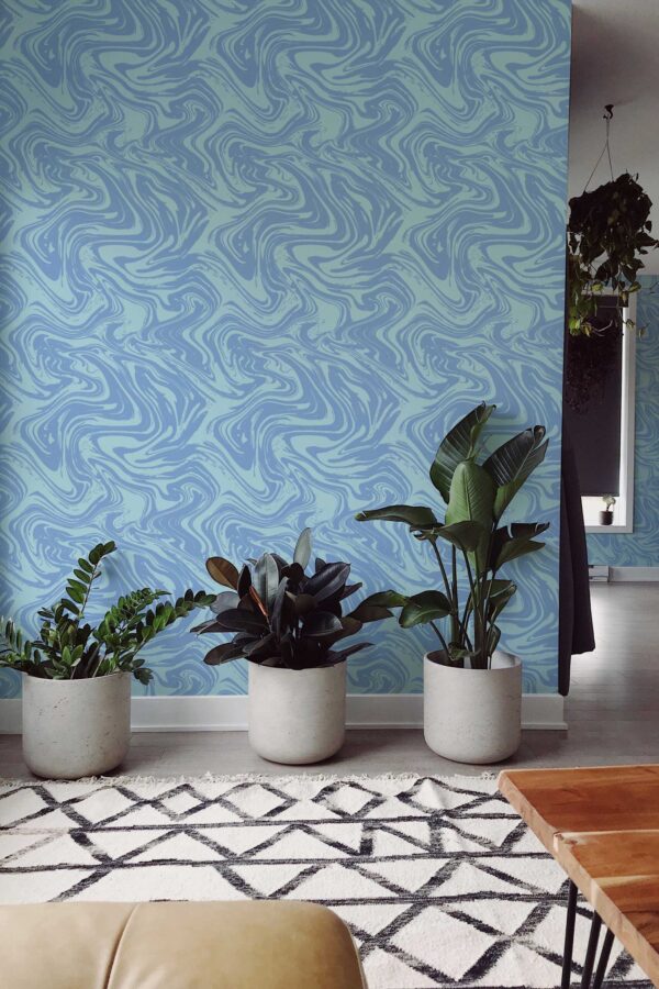A modern living area featuring a swirling blue marble-patterned wallpaper as the accent wall. The wallpaper design shows flowing, water-like swirls in varying shades of sky, aqua, and navy blues, adding a serene, abstract aesthetic. Three large concrete planters filled with lush tropical houseplants sit in front of the wall, complemented by a geometric rug and simple wooden coffee table. The overall style is contemporary and airy, with clean lines and subtle decorative elements that highlight the calming, fluid wallpaper design.