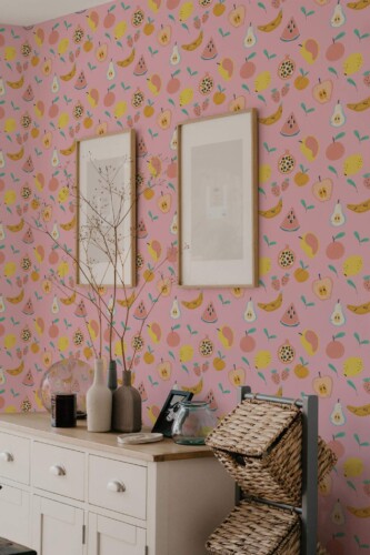 Vibrant kitchen corner showcasing pink fruit wallpaper decorated with scattered yellow bananas, pink pears, red watermelon slices, and white pears. A light wooden sideboard stands in the foreground, topped by muted-gray vases and a translucent glass jar. Two framed artworks hang on the wall, adding subtle contrast against the colorful pattern. A ladder shelf with woven baskets on the right completes this playful, eclectic setting, blending cheerful produce motifs with soft, natural textures.