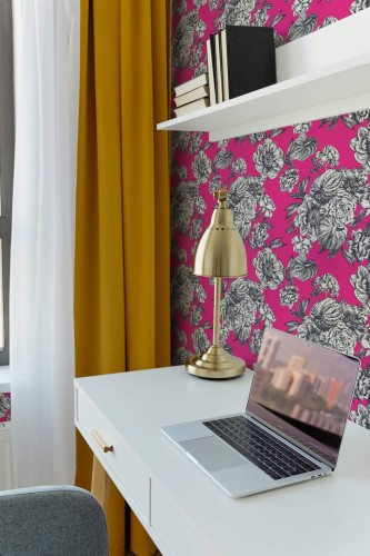 Home office corner featuring hot pink floral wallpaper with bold black-and-white blossoms. A sleek white desk holds an open laptop and a brass desk lamp, adding a touch of metallic shine. Above, a minimalist white shelf displays black-and-white books. Vibrant golden curtains frame a window on the left, creating a striking contrast against the bright pink background. The overall scene feels modern and energizing, blending bold color with refined, practical décor.