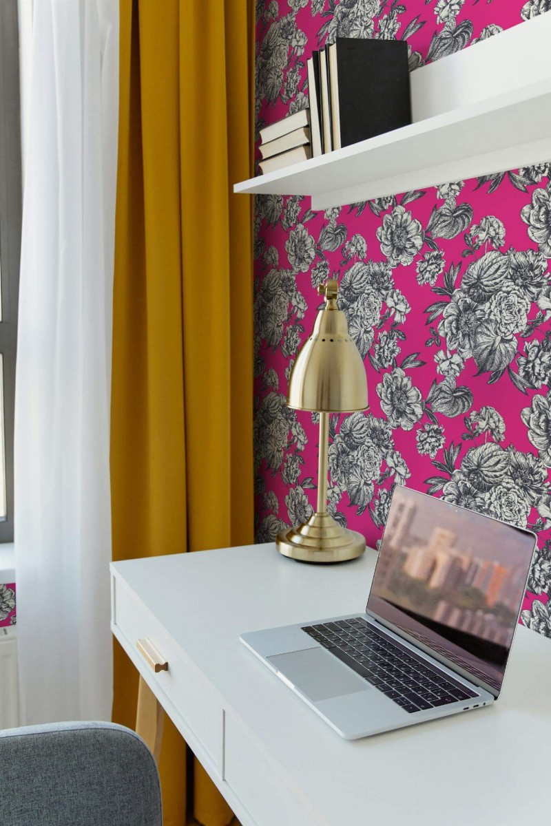 Home office corner featuring hot pink floral wallpaper with bold black-and-white blossoms. A sleek white desk holds an open laptop and a brass desk lamp, adding a touch of metallic shine. Above, a minimalist white shelf displays black-and-white books. Vibrant golden curtains frame a window on the left, creating a striking contrast against the bright pink background. The overall scene feels modern and energizing, blending bold color with refined, practical décor.