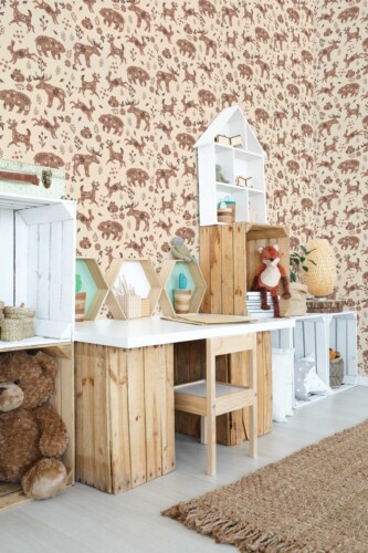 Children’s playroom featuring a whimsical brown woodland animal wallpaper design with Scandinavian-inspired motifs. The wallpaper pattern showcases hand-drawn deer, moose, bears, foxes, and rabbits in soft brown tones on a beige background, with delicate leafy accents creating a forest scene. The room is decorated in a natural, rustic style with a wooden desk made from upcycled crates, cozy storage shelves, a rattan rug, and plush toys. This boho-inspired setting pairs perfectly with the soft color palette and nature-themed pattern, making it ideal for nurseries, play spaces, or reading corners.