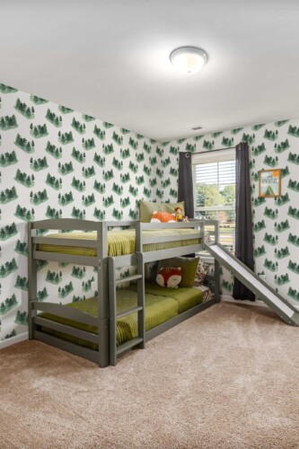 Children’s bedroom featuring a gray bunk bed with green bedding placed against walls covered in a watercolor pine tree wallpaper. The repeating forest pattern, shown in various green shades on a white background, creates a calm, nature-inspired atmosphere. A plush beige carpet adds warmth underfoot, while a window draped with dark curtains allows natural light to brighten the space. Plush toys rest on the upper bunk, and a small decorative sign near the window adds a playful finishing touch.
