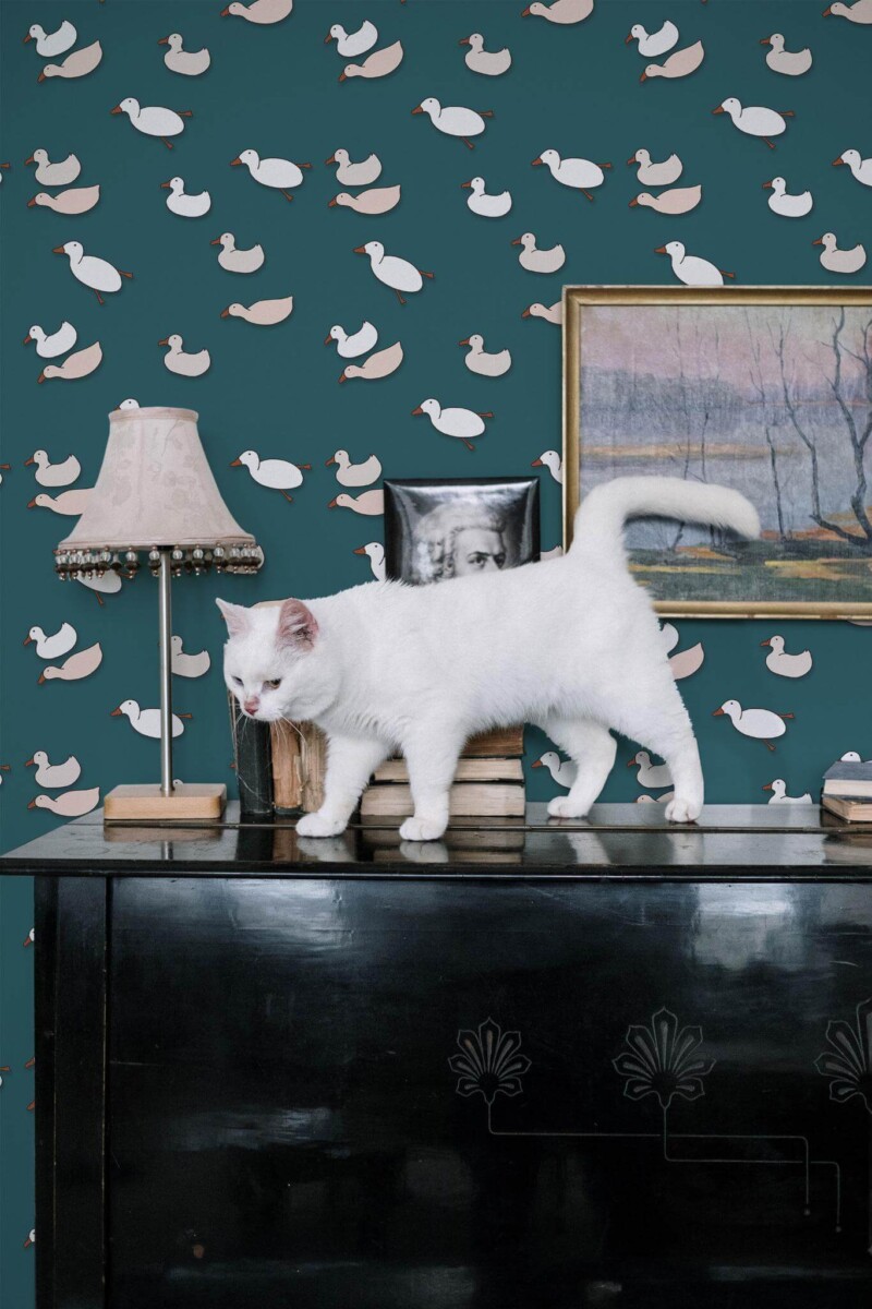 Whimsical accent wall decorated with teal cartoon duck wallpaper featuring scattered white-and-pink ducks. A glossy black piano sits in front, topped by vintage books, a small framed painting, and an antique table lamp with a patterned shade. A curious white cat walks across the books, drawing attention to the playful, eclectic décor. The wallpaper’s imaginative duck motif contrasts with the cat’s bright fur, creating a unique and visually engaging scene.