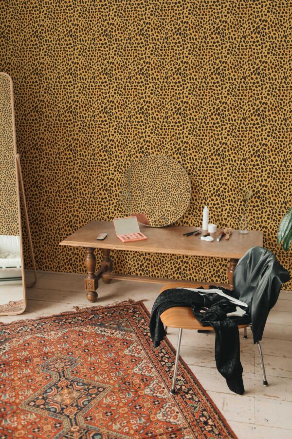 Eclectic interior showcasing tiny cheetah print wallpaper in golden yellow with bold black spots. A richly patterned rug in warm reds and oranges covers the hardwood floor, adding depth. A vintage wooden desk, topped by a round woven tray and slender white candles, anchors the center. A sleek mid-century chair with a black jacket draped over it sits nearby. On the left, a tall mirror reflects natural light, enhancing the room’s dramatic yet cozy ambiance.