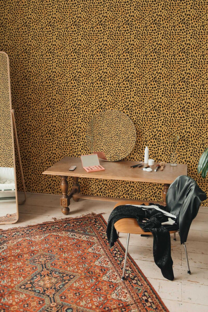 Eclectic interior showcasing tiny cheetah print wallpaper in golden yellow with bold black spots. A richly patterned rug in warm reds and oranges covers the hardwood floor, adding depth. A vintage wooden desk, topped by a round woven tray and slender white candles, anchors the center. A sleek mid-century chair with a black jacket draped over it sits nearby. On the left, a tall mirror reflects natural light, enhancing the room’s dramatic yet cozy ambiance.