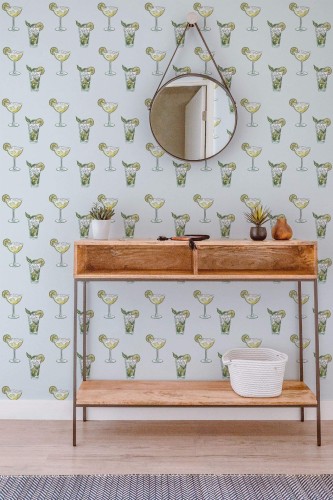 Entryway featuring a light-blue cocktail wallpaper with repeating margarita and mojito glass illustrations in soft green and yellow tones. A round mirror hangs above a rustic wooden console table with slim metal legs, providing a modern yet earthy accent. Potted succulents and small decorative items rest on the table’s surface, adding natural texture. The light wood flooring and subtle woven rug complete the airy, whimsical vibe, merging playful bar-inspired décor with fresh, minimalist styling.