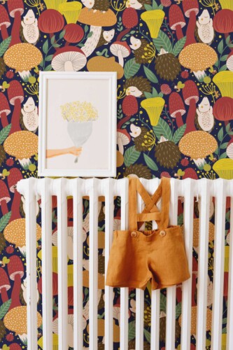 A nursery scene featuring whimsical wallpaper with playful hedgehogs and colorful mushrooms against a deep navy background. A framed floral print rests above a white radiator adorned with burnt orange toddler suspenders, tying together the bright reds, yellows, and earthy neutrals. The overall style blends a nature-inspired woodland theme with modern, minimalist touches. This cozy and vibrant interior highlights the wallpaper’s fun, child-friendly design, creating an inviting space perfect for imaginative play