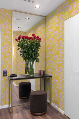 A vibrant, eclectic interior featuring Fancy Walls “Sunset Wildcat Print” wallpaper, designed with a bold animal-inspired pattern in soft lavender and golden orange brushstroke spots. The wallpaper adds a dynamic, energetic feel to the space, creating a lively backdrop in a modern vanity area. A sleek black and gold console table with a round velvet stool and a tall mirror enhances the chic styling, while a vase of fresh red roses adds a romantic contrast. Available as peel and stick or non-pasted wallpaper, this design is printed with GreenGuard Gold certified inks on eco-conscious, PVC-free materials—perfect for stylish, personality-filled spaces.