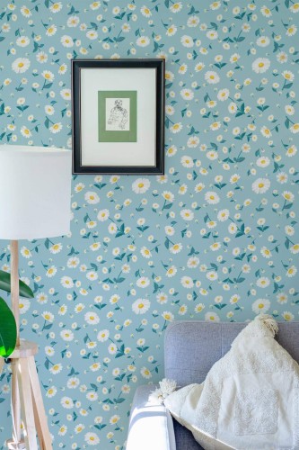 Living room corner featuring a light blue daisy wallpaper with scattered white-and-yellow daisies and teal leaves. A gray armchair sits on the right, topped by a neutral throw blanket. In the center, a small framed art print hangs on the wall, providing a subtle contrast against the cheerful floral pattern. A wooden floor lamp with a white lampshade stands to the left, adding a warm, cozy element that complements the bright, nature-inspired design.
