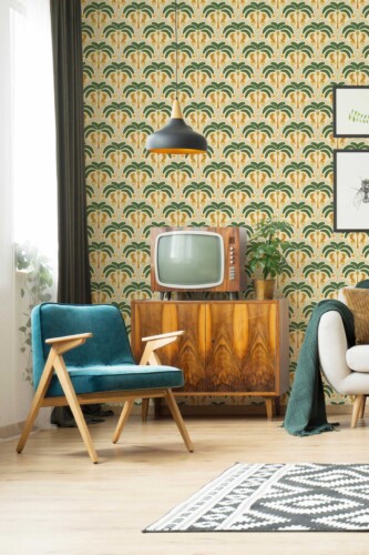 A retro-inspired living room featuring a bold Art Deco Jungle wallpaper with repeating symmetrical patterns of palm trees and golden leopards on a warm beige background. The palm fronds are deep green with detailed linear textures, while the leopards face each other beneath each tree, creating mirrored focal points across the wall. The pattern is accented with small yellow flowers and dotted arches in orange and green, forming a rhythmic, vintage aesthetic. In the foreground, a mid-century modern wood cabinet with rich grain patterns supports a vintage television. A teal velvet armchair with natural wood arms and legs sits nearby, adding a pop of color and texture.