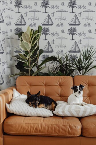 Hand-drawn coastal-themed wallpaper with navy blue illustrations of sailboats, palm trees, sun umbrellas, beach chairs, bicycles, and beach balls, repeated on a soft white background. The wallpaper adds a vintage seaside charm to the space, creating a relaxed and breezy atmosphere. Shown in a cozy living room with lush green plants, a camel leather sofa, and two small dogs lounging on striped pillows. The Fancy Walls peel and stick or non-pasted wallpaper brings warmth and personality to any beach-inspired or eclectic interior.