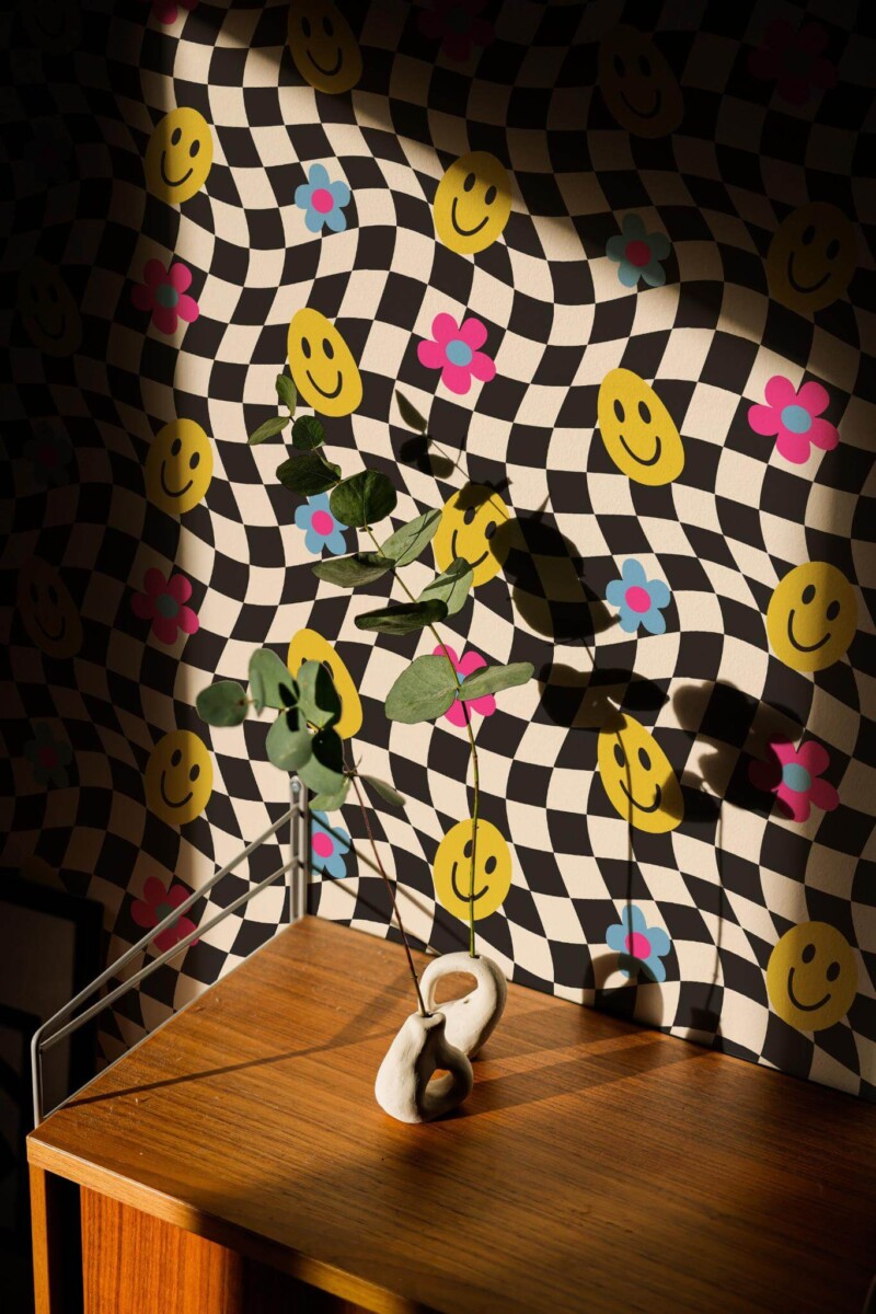 Funky Wallpaper with a wavy black-and-white checkered background, decorated by bright yellow smiley faces and pink flowers, styled in a sunlit corner with a wooden side table and minimalist vase of eucalyptus. The bold, playful pattern adds a lively, modern accent to the room.
