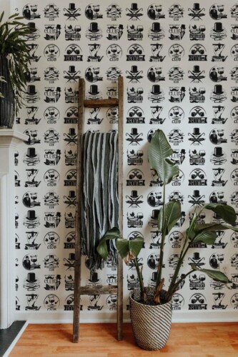 Black and white Gentlemen’s Club wallpaper featuring vintage-inspired motifs like top hats and refined emblems. Displayed in a cozy interior with a rustic ladder, woven basket, and lush green plant for a sophisticated, eclectic look.