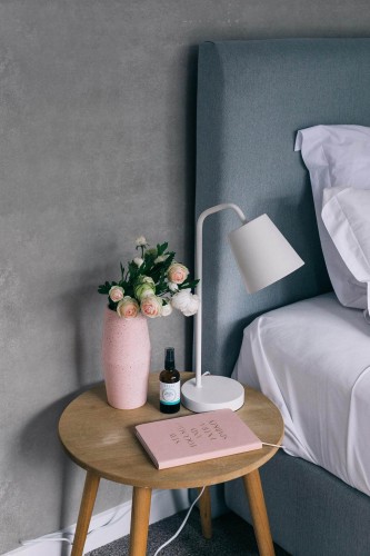 This serene and minimalist bedroom features a soft gray concrete wallpaper with subtle texture, creating a calm and sophisticated backdrop. The design adds depth without overpowering the space, making it an ideal choice for creating a tranquil atmosphere. A bedside table with a simple wooden design is adorned with a pink vase holding fresh flowers, a small bottle of essential oil, and a pink notebook, completing the gentle, cohesive aesthetic. The Fancy Walls peel and stick or non-pasted wallpaper creates a refined ambiance for any bedroom, with easy installation and eco-friendly materials.