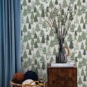 Evergreen Christmas Tree Wallpaper with stylized pine trees in varying shades of green on a light background, styled in a cozy space with a wooden side table, a teal curtain, and a wicker basket filled with colorful yarn. The festive forest motif adds warm, seasonal charm to the room.