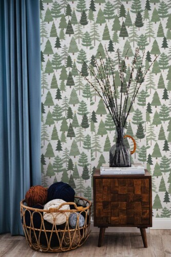 Evergreen Christmas Tree Wallpaper with stylized pine trees in varying shades of green on a light background, styled in a cozy space with a wooden side table, a teal curtain, and a wicker basket filled with colorful yarn. The festive forest motif adds warm, seasonal charm to the room.