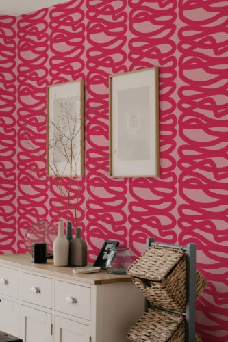 Hot pink line wallpaper featuring bold, freehand “spaghetti” loops on a lighter pink background. Displayed in a living space with a wooden sideboard, decorative vases, framed prints, and woven baskets, creating a vibrant, modern statement wall.