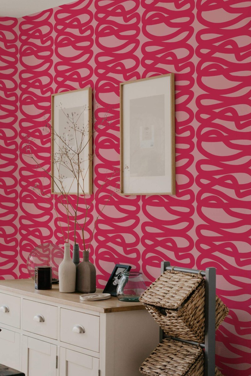 Hot pink line wallpaper featuring bold, freehand “spaghetti” loops on a lighter pink background. Displayed in a living space with a wooden sideboard, decorative vases, framed prints, and woven baskets, creating a vibrant, modern statement wall.