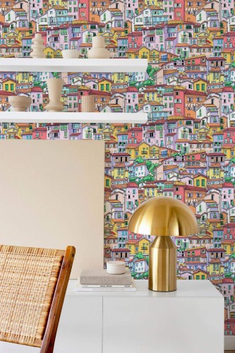 Modern style dining room decorated with Italy peel and stick wallpaper featuring a vibrant, hand-drawn illustration of colorful hillside houses inspired by Italian coastal towns. The playful mural is paired with minimalist white shelving, natural wood textures, a sculptural golden lamp, and neutral-toned ceramics. This eclectic yet balanced interior blends contemporary design with Mediterranean charm and creative wall décor.